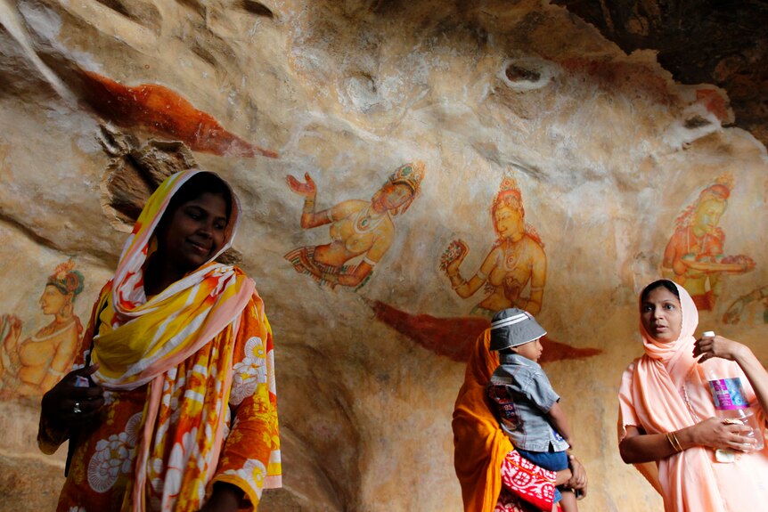Women look at orange, red and yellow frescoes of ancient drawn figurines on a rock.