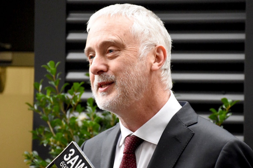 A man in a suit and tie speaks into a microphone at an outdoor press stop.