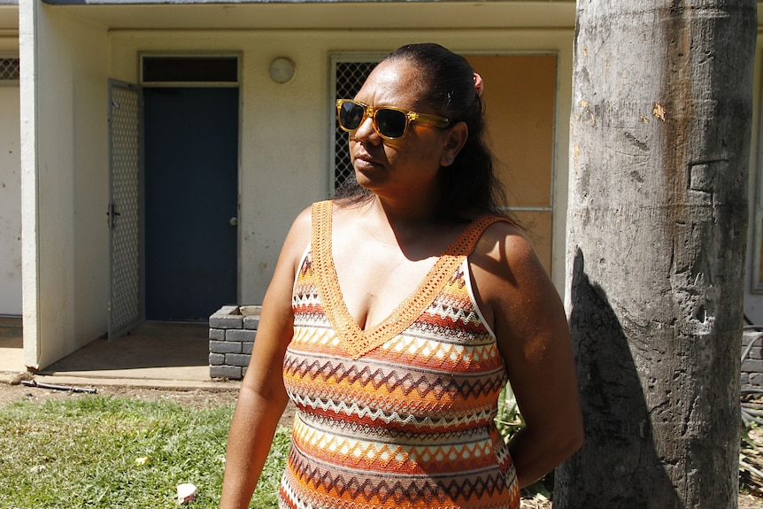 A woman in sunglasses stands in front of a residential building looking out.