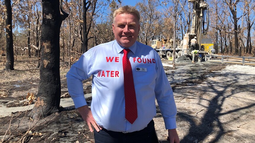 A man stands in a burnt out forest with a shirt that says 'we found water'