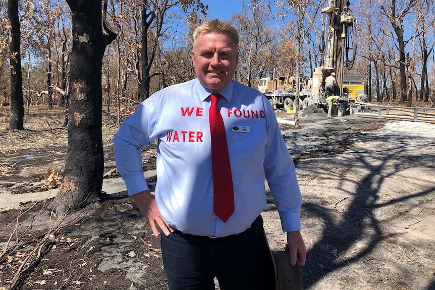 A man stands in a burnt out forest with a shirt that says 'we found water'