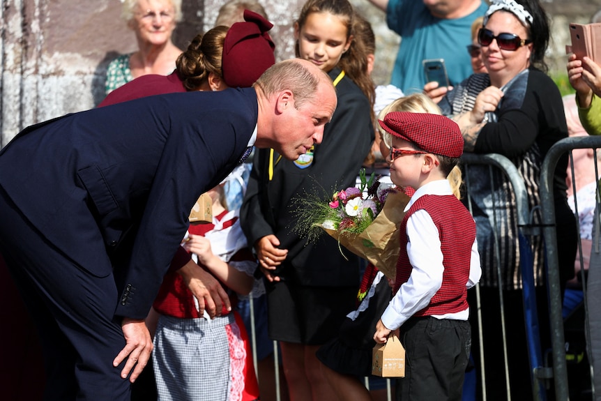 Prince William wearing a long coat bends down to talk to a fan wearing a cap and glasses.