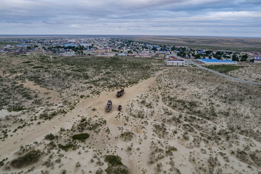An aerial view of the dried out Aral sea, sandy ground with rusty ships, a town lies in the distance. 
