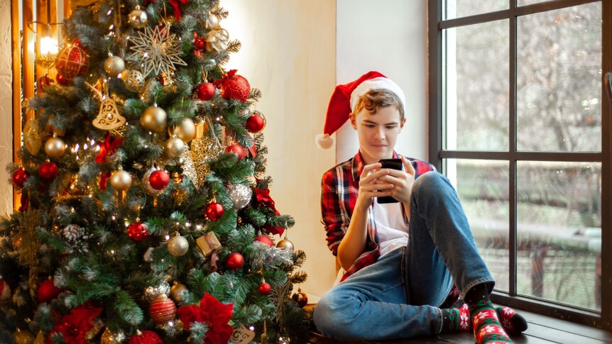 Teenage boy wearing Santa hat holds mobile phone and sits on windowsill next to Christmas tree