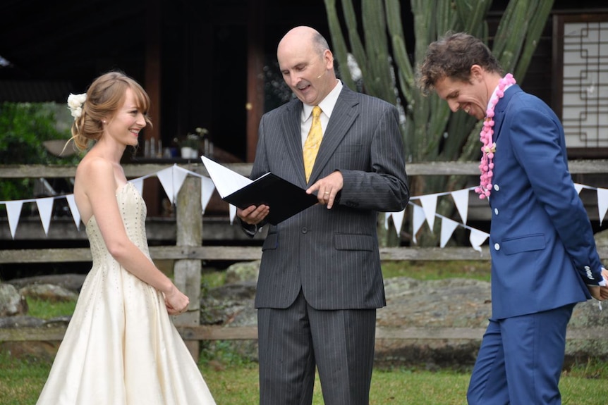 Pic of Shaun Wood officiating outdoors as a celebrant, standing between a bride and a groom.