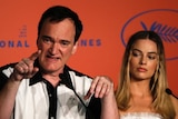 Tarantino speaks into the microphone, pointing, while seated next to Robbie