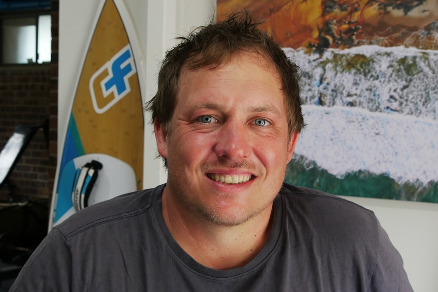 A man with blond brown hair smiles, there is a surfboard and a beach photo in the background.