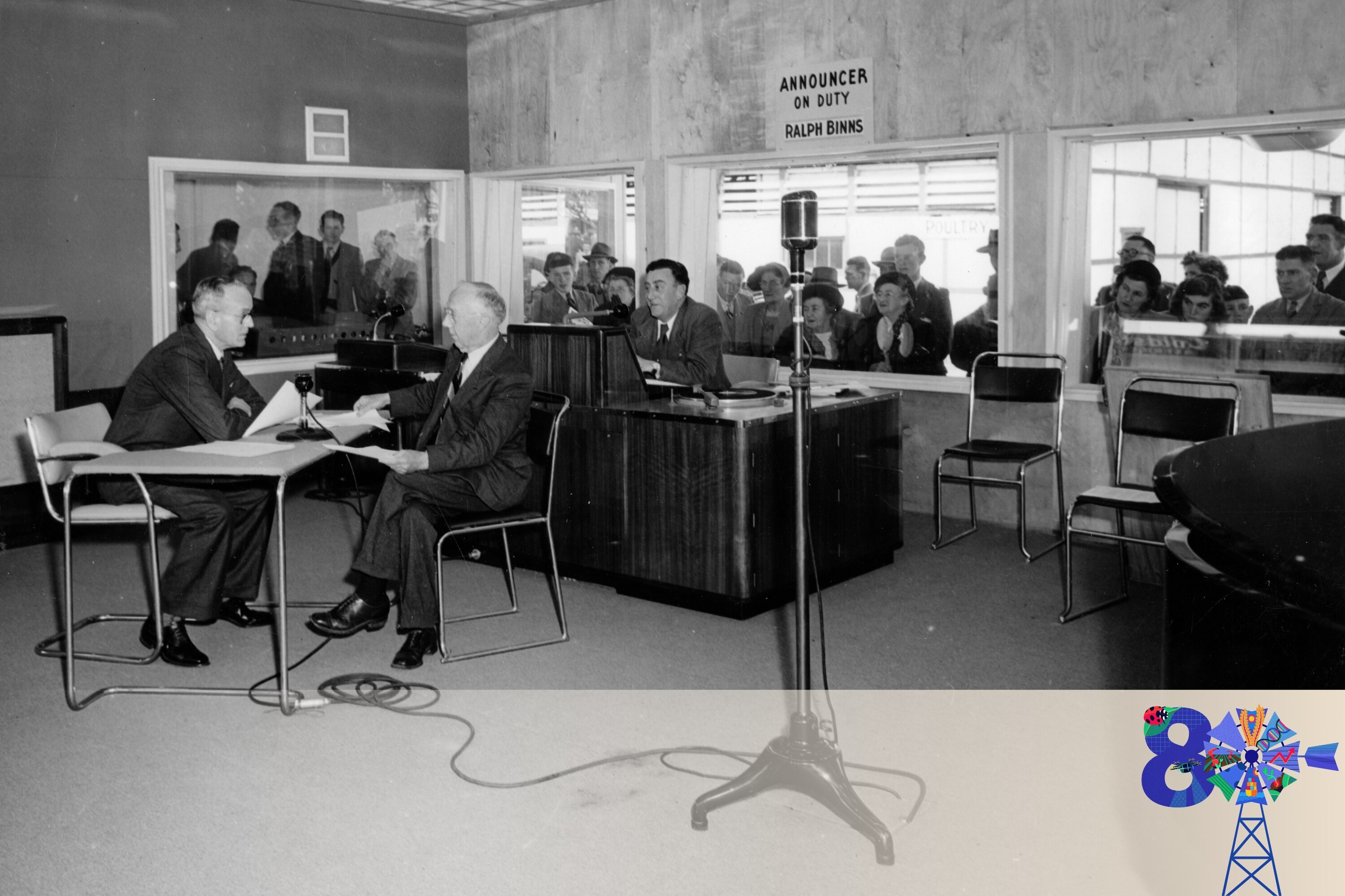 Black and white image of two men sitting at a desk presenting a radio program while onlookers observe through windows 
