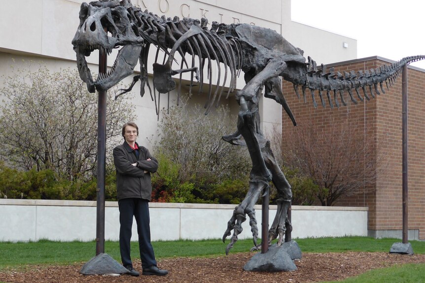 A young man stand beneath a replica skeleton of a Tyrannosaurus Rex.