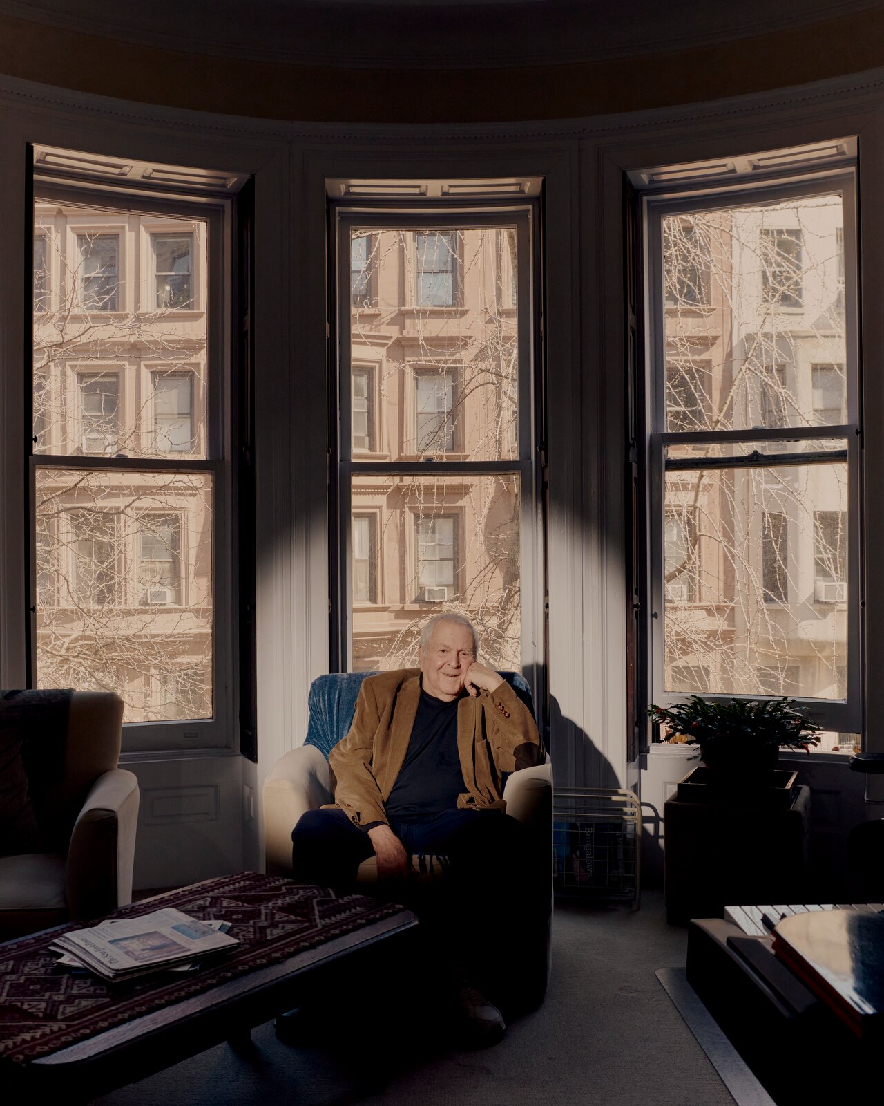 An elderly white man in a dark shirt and tan jacket sits in his New York apartment in an armchair in front of a bay window.