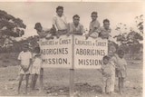 A group of 11 boys poses around a sign reading 'Churches of Christ Aborigine Mission'. The photograph is black and white.
