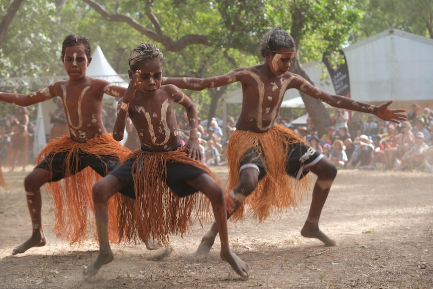 A group of boys and men wearing body paint and traditional Aboriginal dress perform a dance.