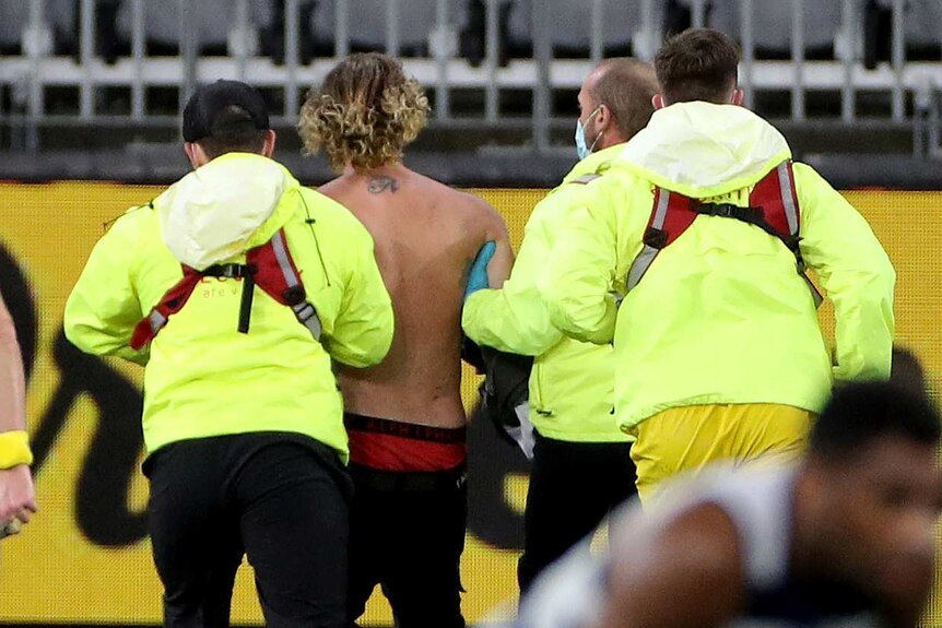 Four secuity guards in yellow hi-vis jackets and face masks escort a pitch invader off Perth Stadium with an umpire nearby.