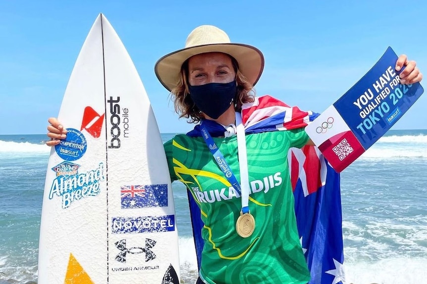 Woman standing wearing face mask holding a surf board and a large ticket saying she has qualified for the OIympics