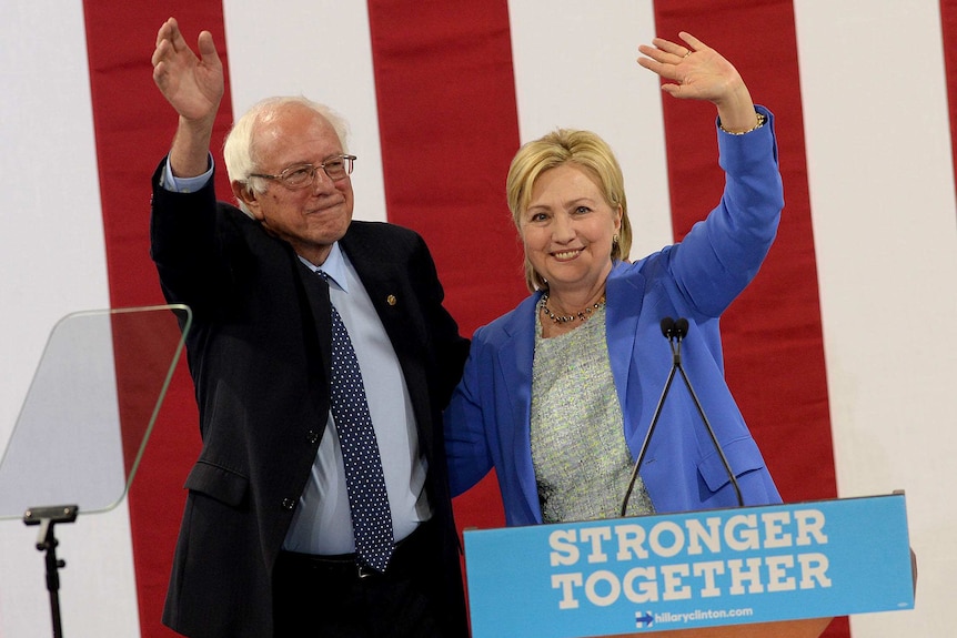 Bernie Sanders and Hillary Clinton embrace while waving to a crowd.