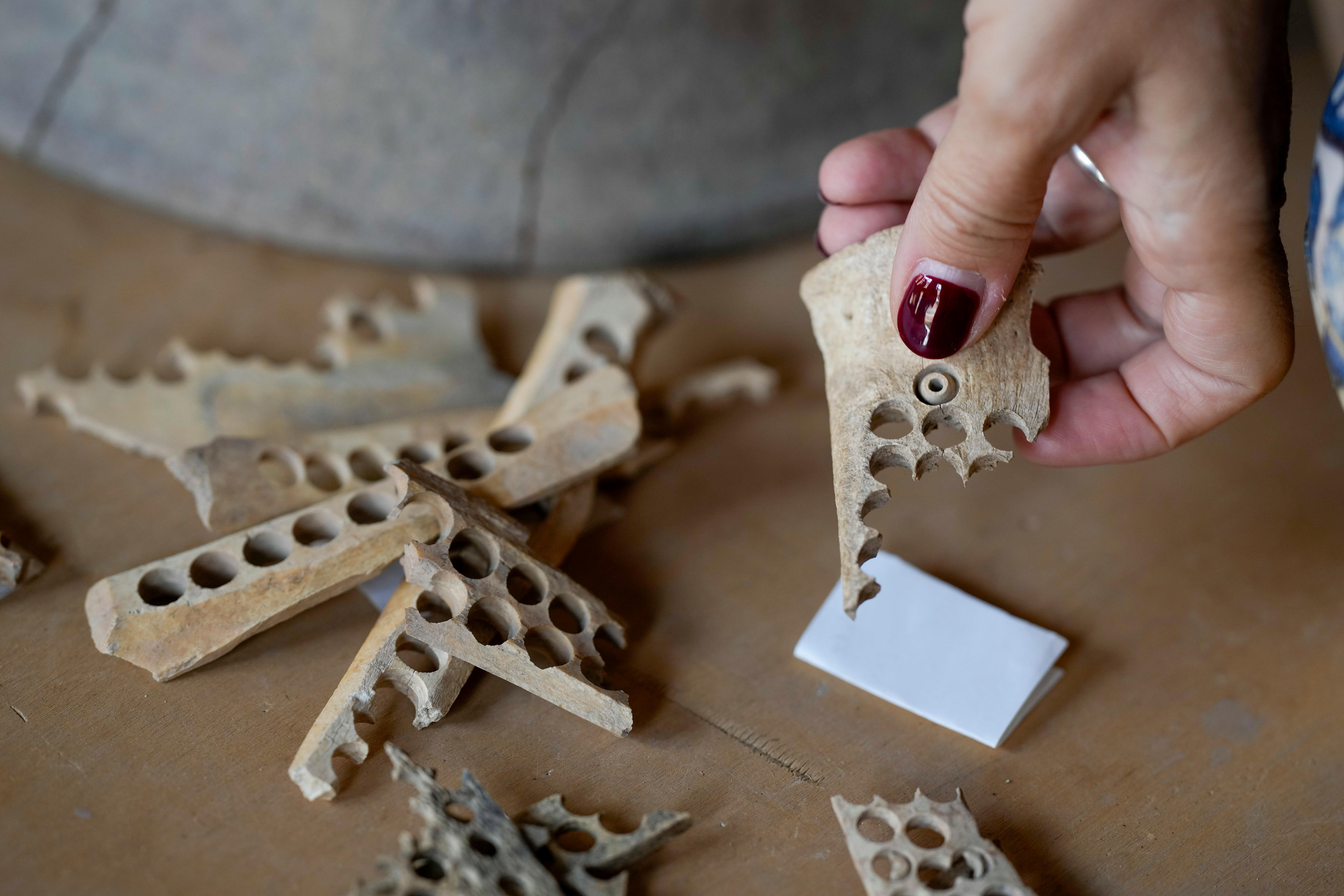 a hand holding an ancient broken piece of bone with circle holes punctured through and several others on a table below 