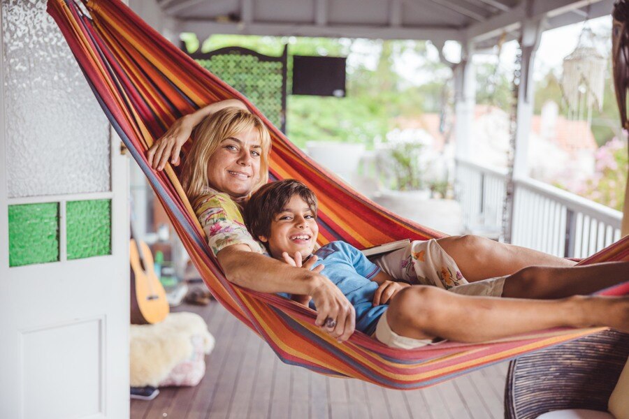 A blonde woman sits in a hammock with her young son, on the front deck of her house, smiling at the camera.