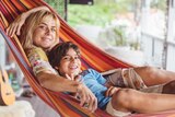A blonde woman sits in a hammock with her young son, on the front deck of her house, smiling at the camera.