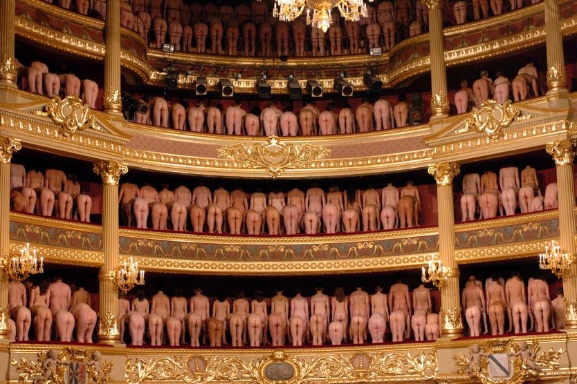 Naked models stand, with their backs turned, on several levels of theatre seating.