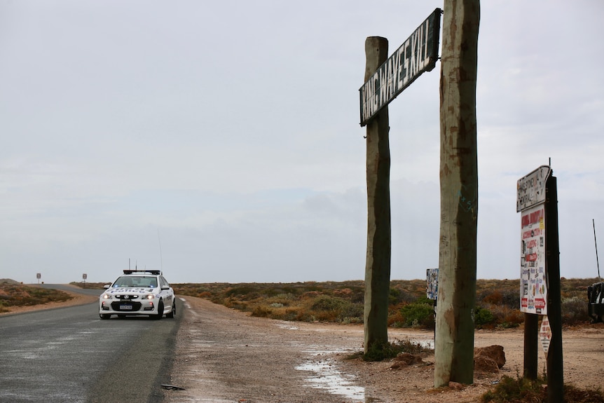 A police car on a ountry road with a large wooden sign off to the right of the road.