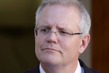 A man with glasses and an Australian flag pin on his jacket looks into the distance