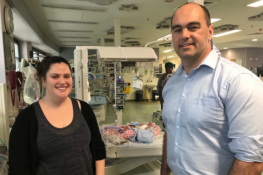 A woman, man and baby in a cot in a hospital setting.