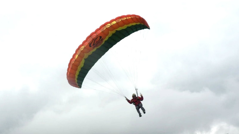 A man suspended below a parachute comes in to land on a grassy field in front of mountains.