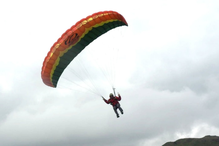 A man suspended below a parachute comes in to land on a grassy field in front of mountains.