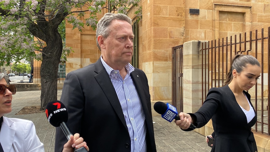 Reporters point microphones at a man wearing a suit and a blue stripy shirt outside a stone building