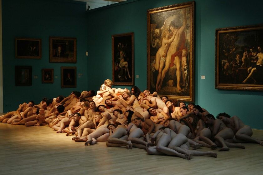 Naked people are stacked on top of one another in front of framed paintings in a museum.