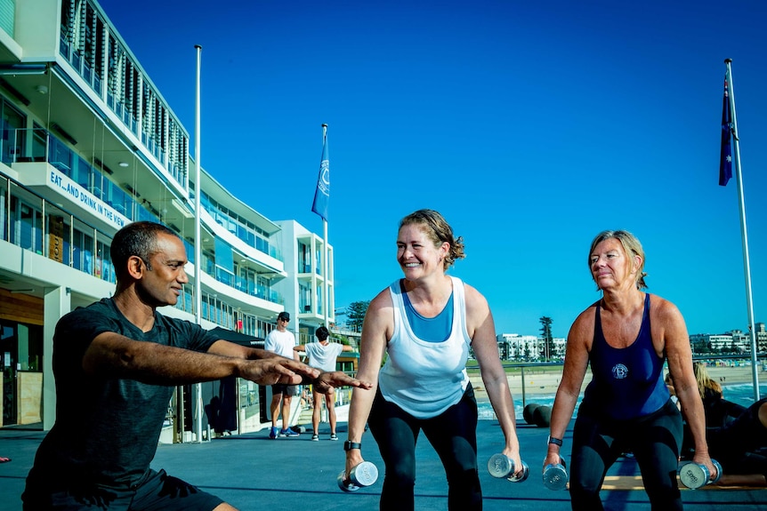 A man demonstrates a squat to two women at an outdoor gym