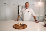 A man in a chef's outfit stands smiling in a kitchen with small desserts on a serving platter on a bench in front of him.