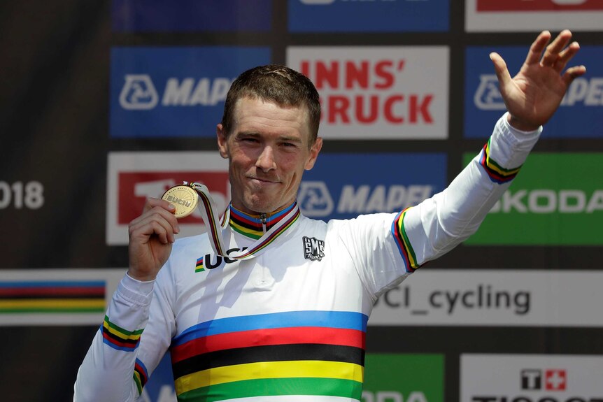Rohan Dennis stands on a podium waving and holding his medal