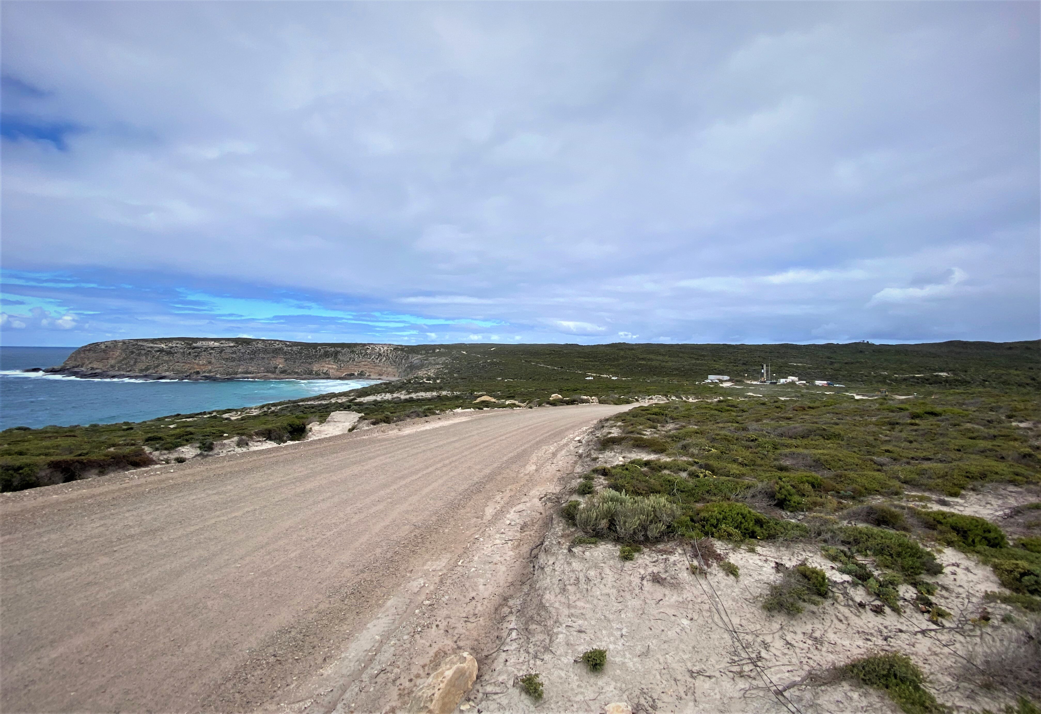Coastal scene with road in foreground, ocean and headland to left, and green on right, with rocket, buildings seen in distance