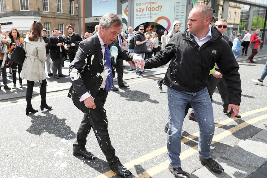 A man in a suit covered with milkshake is led away by another man holding his arm.