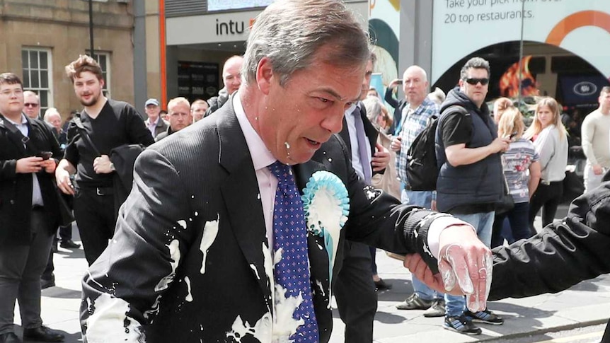A man in a suit covered with milkshake is led away by another man holding his arm.