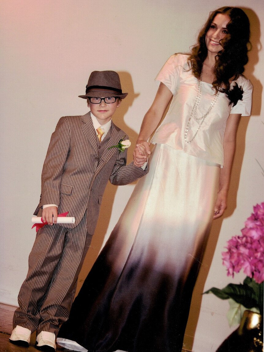 A young Connor O'Grady, an aspiring fashion designer in Brisbane, dressed in a suit and hat holds hands with a woman.