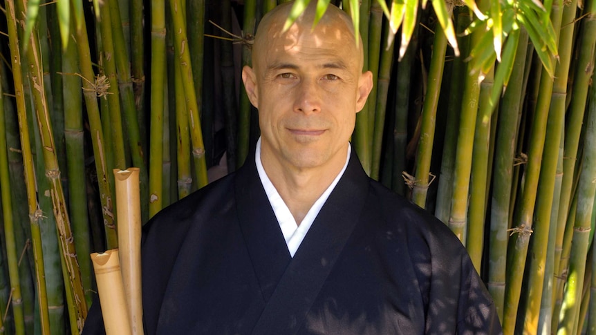 A man stands under bamboo holding two Japanese bamboo flutes.