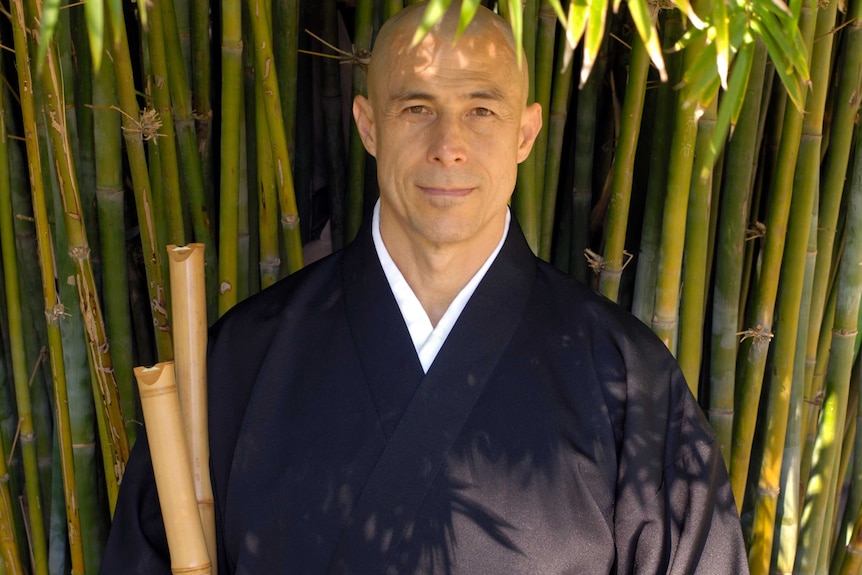 A man stands under bamboo holding two Japanese bamboo flutes.