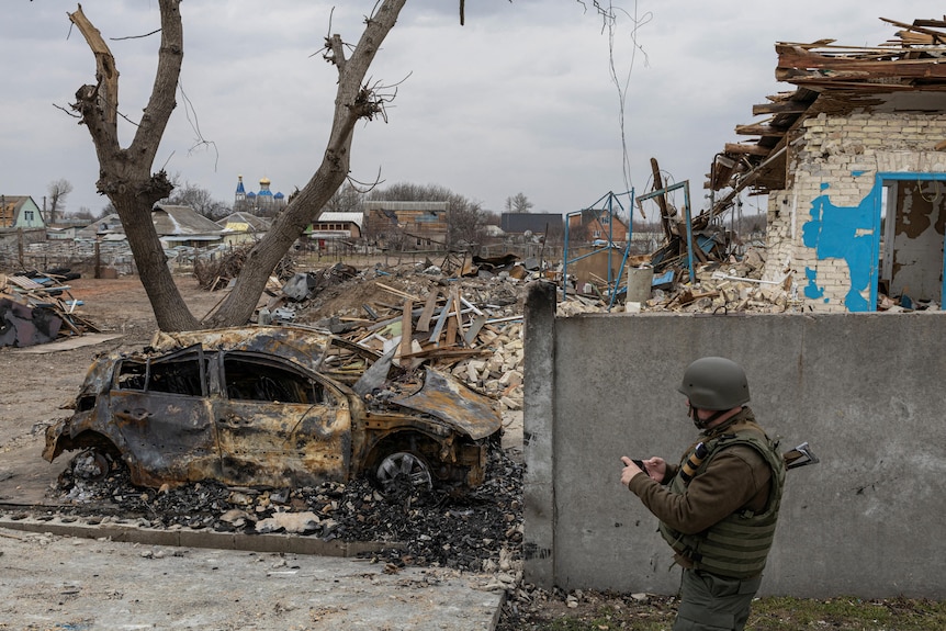A Ukrainian serviceman walks past a burnt-out car and damaged house.