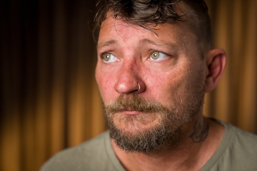 A man with a goatee, short hair and a nose piercing looks upwards with a sad expression