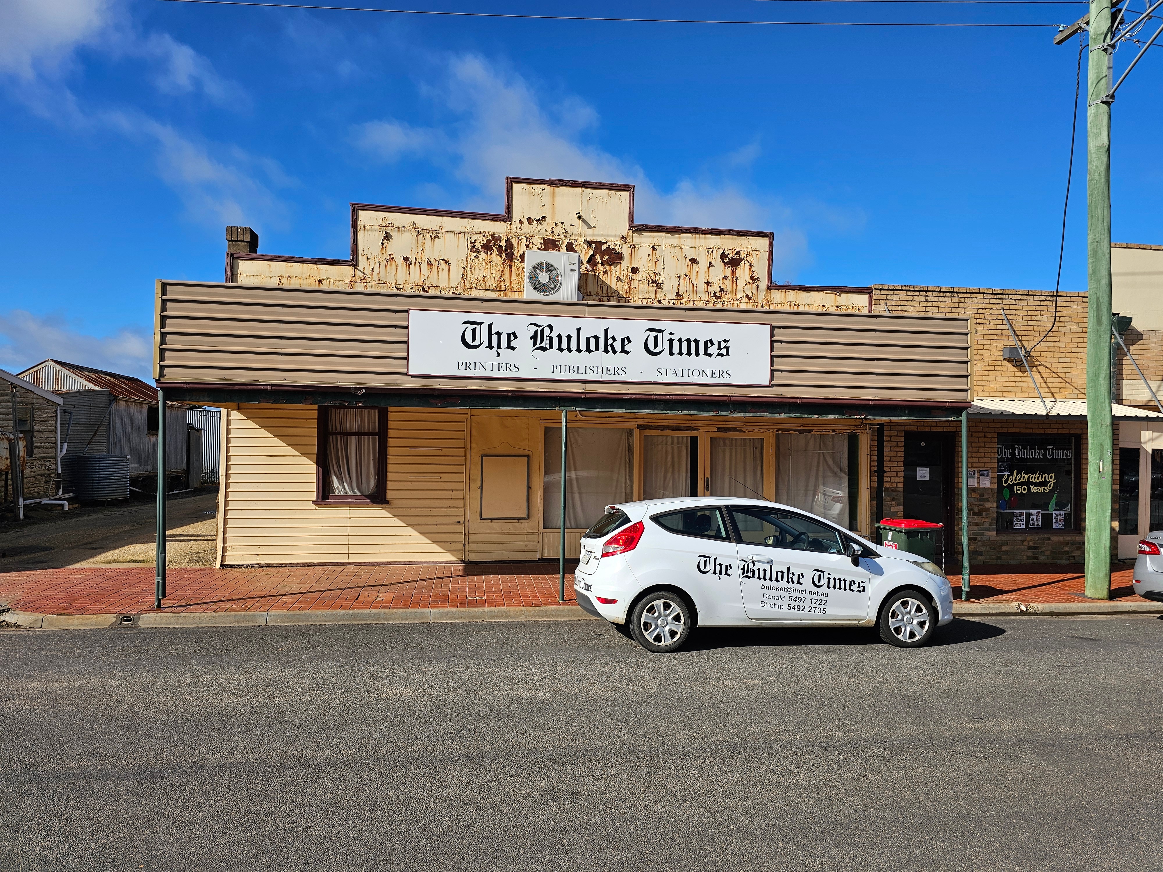 Cream-coloured Victorian shopfront with  Buloke times masthead on its front. Blue sky, country road, a car with name in front.