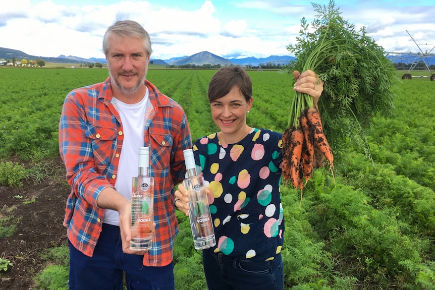 Jason Hannay and Gen Windley standing in a carrot farm holding carrots and a bottle of carrot vodka.
