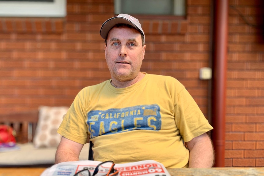 A man in a yellow t-shirt and grey cap seated at a table, with a newspaper, iPhone and glasses in front of him.