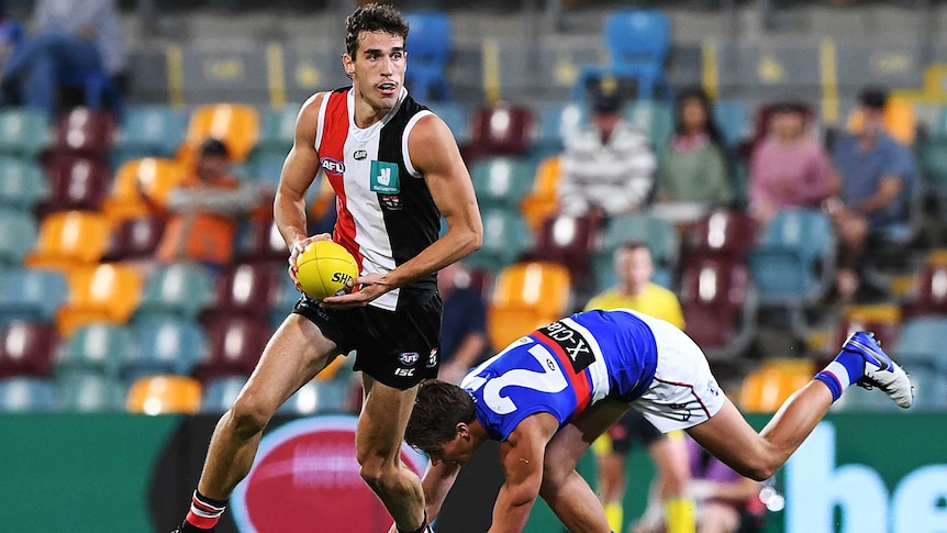 A tall AFL forward looks up and runs with the ball as a defender stumbles after missing a tackle.