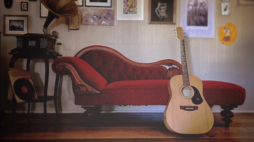 A guitar leaning against a red couch, in front of a wall with many framed pictures on it.