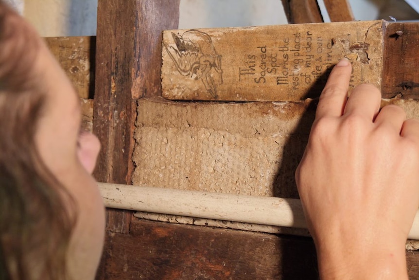 A woman in her 30s looks closely at the writing on a piece of cross embedded as part of the house wall.