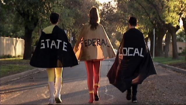 Rear view of teenagers walk in street wearing capes, capes have text: "State", "Federal", "Local"