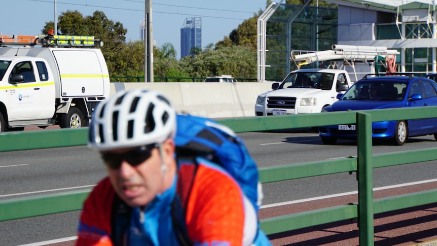 Cyclist in foreground riding along pathway, alongside freeway with cars and city in the background.