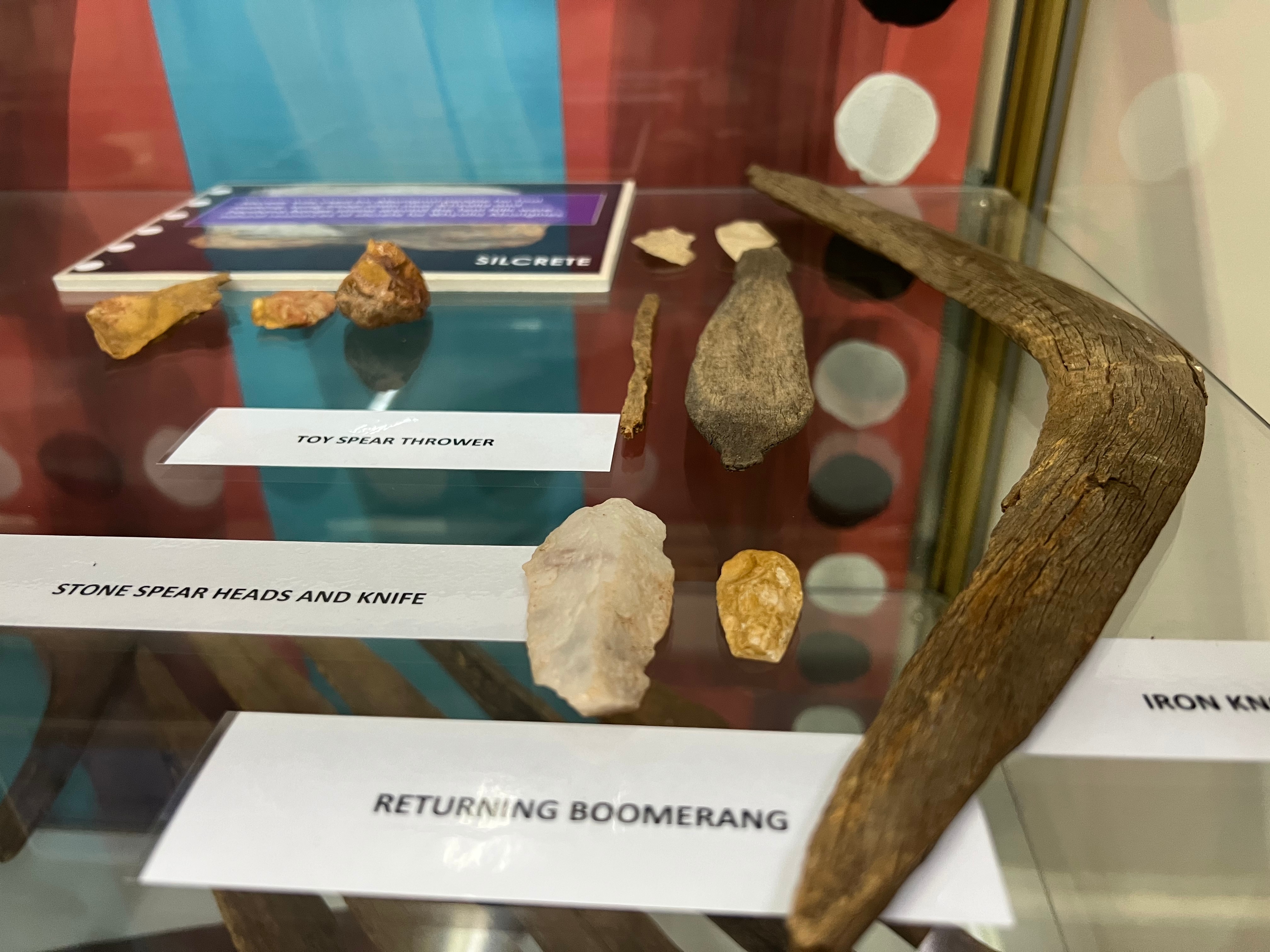 A series of ancient indigenous tools, including a toy spear launcher, in a glass cabinet. 